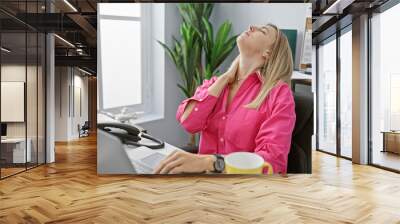 A young, blonde woman in a pink shirt feeling neck pain at her office desk, exuding a mix of professionalism and discomfort. Wall mural