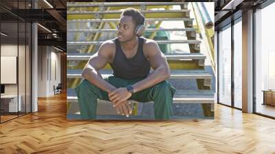 A young african american man with headphones sits reflectively on urban stairs outdoors. Wall mural