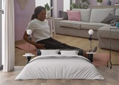 A young african american woman with curly hair is sitting on a mat, exercising in a cozy living room of her home, showcasing a calm and relaxed indoor setting. Wall mural