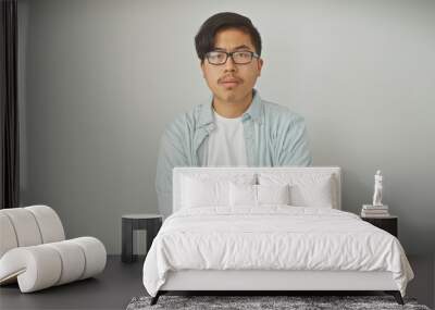 A young asian man in casual attire stands with crossed arms against a white background, exuding confidence. Wall mural