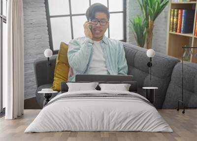 A young asian man multitasks with a phone call and laptop in a modern living room. Wall mural