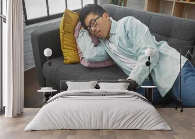 A young asian man sleeps on a couch with colorful pillows in a cozy apartment living room, remote in hand. Wall mural