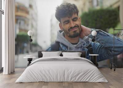 A young bearded hispanic man in casual attire on a city street, expressing neck pain. Wall mural