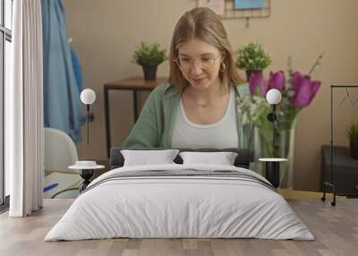 A young blonde woman wearing glasses works on her laptop at home, surrounded by plants and flowers. Wall mural