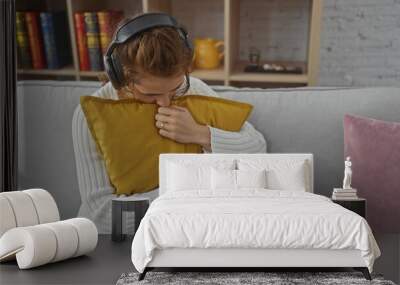 A young caucasian woman in casual attire is relaxing at home, hugging a pillow on the sofa with headphones. Wall mural