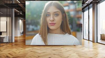 A young caucasian woman poses outdoors in a city, exuding natural beauty with her casual attire and warm gaze Wall mural