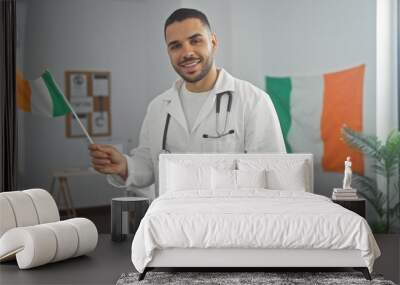 A young hispanic male doctor smiling, holding an irish flag indoors with a stethoscope around his neck. Wall mural