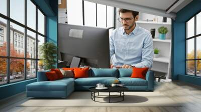 A young hispanic man in a blue shirt working attentively at his office desk indoors, illustrating a professional business environment. Wall mural