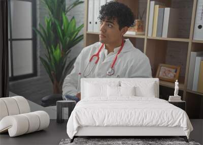 A young hispanic man in a white coat uses a smartphone in a hospital office, appearing thoughtful and professional. Wall mural