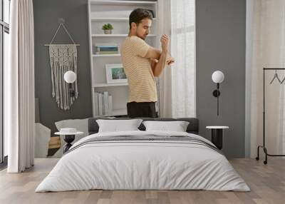 A young hispanic man in casual attire is standing thoughtfully in a well-lit, modern bedroom with stylish decor. Wall mural