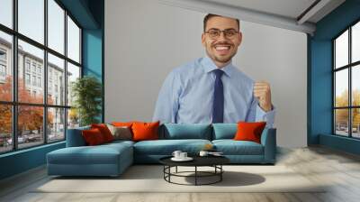 A young hispanic man with a beard and glasses in a blue shirt and tie stands isolated against a white background with a confident smile and clenched fist. Wall mural