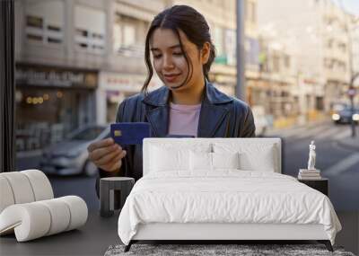 A young hispanic woman examines her credit card while holding a smartphone on a sunny city street. Wall mural