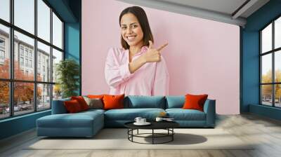 A young hispanic woman in a pink shirt points to the side against a plain pink background, exuding confidence. Wall mural