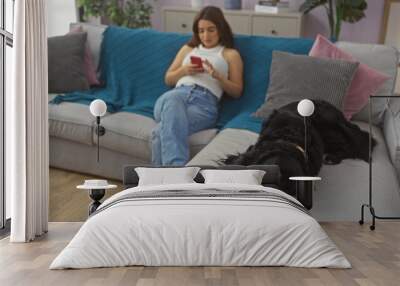 A young hispanic woman in casual attire sits on a living room sofa looking at her phone, while a black labrador dog lies next to her chewing on a toy inside a cozy apartment. Wall mural