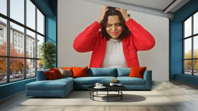 A young hispanic woman looking frustrated, with hands on her head, isolated against a white wall background wearing a red cardigan and white shirt. Wall mural