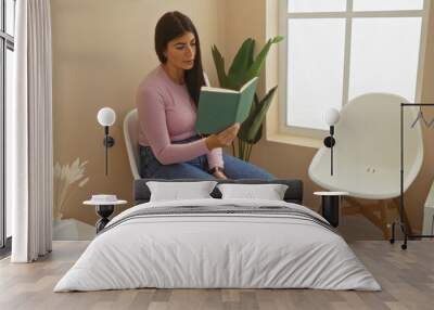 A young hispanic woman reading a book while sitting in a modern indoor waiting room with natural light and minimalist decor. Wall mural