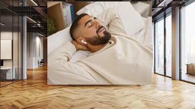 A young man with a beard wearing a wireless earphone naps peacefully on a white bed in a cozy bedroom setting. Wall mural