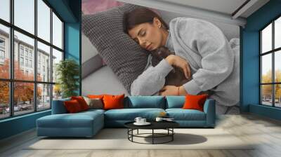 A young woman in casual attire affectionately embraces her sleeping pet chihuahua on a comfortable sofa indoors. Wall mural