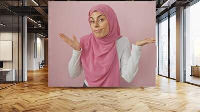 A young woman wearing a hijab poses with a puzzled expression against a pink background. Wall mural