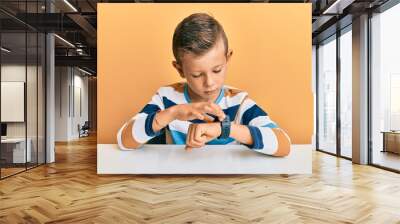 Adorable caucasian kid wearing casual clothes sitting on the table checking the time on wrist watch, relaxed and confident Wall mural