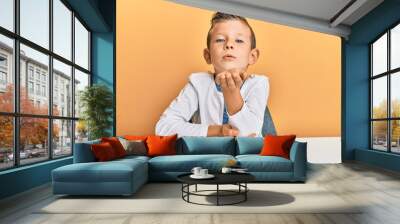 Adorable caucasian kid wearing casual clothes sitting on the table looking at the camera blowing a kiss with hand on air being lovely and sexy. love expression. Wall mural