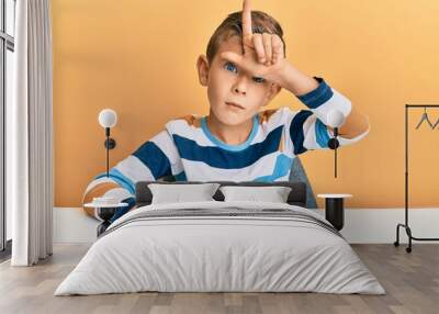 Adorable caucasian kid wearing casual clothes sitting on the table making fun of people with fingers on forehead doing loser gesture mocking and insulting. Wall mural