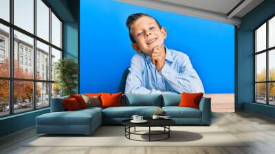 Adorable caucasian kid wearing casual clothes sitting on the table smiling looking confident at the camera with crossed arms and hand on chin. thinking positive. Wall mural