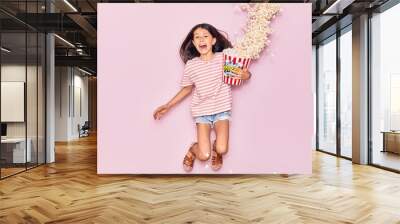 Adorable hispanic child girl surprised with open mouth holding bucket of popcorn jumping over isolated pink background Wall mural