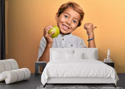 Adorable latin kid holding green apple pointing thumb up to the side smiling happy with open mouth Wall mural