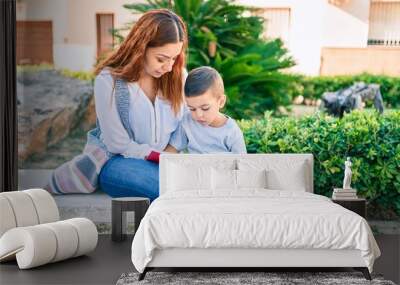 Adorable latin mother and son sitting on the bench and reading book at the park. Wall mural