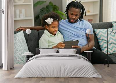 African american father and daughter using touchpad sitting on sofa at home Wall mural