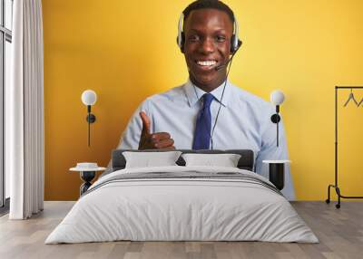 African american operator man working using headset over isolated yellow background doing happy thumbs up gesture with hand. Approving expression looking at the camera with showing success. Wall mural