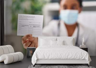 African american woman doctor wearing medical mask holding covid-19 vaccination record card at clinic Wall mural