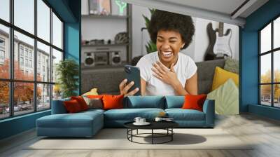 African-american woman laughs while looking at smartphone in cozy living room interior. Wall mural