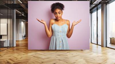 African american woman wearing casual striped dress standing over isolated pink background clueless and confused expression with arms and hands raised. Doubt concept. Wall mural