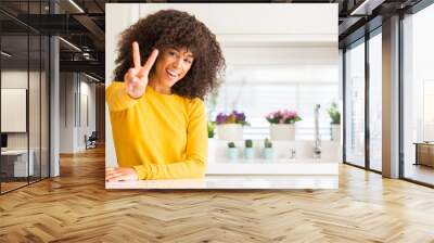 African american woman wearing yellow sweater at kitchen smiling looking to the camera showing fingers doing victory sign. Number two. Wall mural