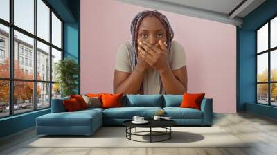 African american woman with braids wearing casual attire posing against a pink background, covering mouth with hands in surprise. Wall mural