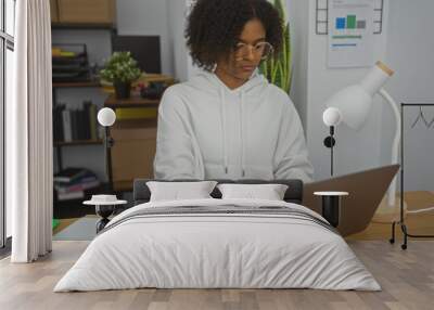 African american woman with curly hair works on a laptop in a modern office setting, wearing a white hoodie and glasses, emphasizing a professional indoor work environment. Wall mural