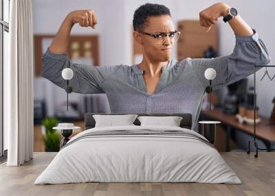 African american woman working at the office wearing glasses showing arms muscles smiling proud. fitness concept. Wall mural