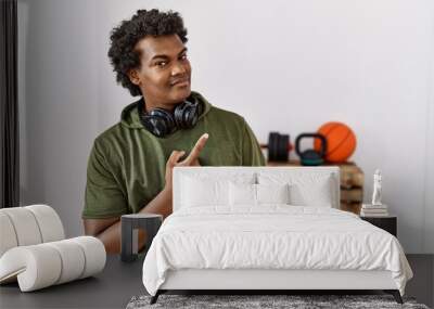 African man with curly hair wearing sportswear at the gym cheerful with a smile of face pointing with hand and finger up to the side with happy and natural expression on face Wall mural