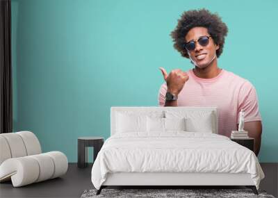 Afro american man wearing sunglasses over isolated background smiling with happy face looking and pointing to the side with thumb up. Wall mural