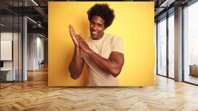 American man with afro hair wearing striped t-shirt standing over isolated yellow background clapping and applauding happy and joyful, smiling proud hands together Wall mural