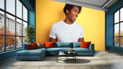 American man with afro hair wearing white t-shirt standing over isolated yellow background looking away to side with smile on face, natural expression. Laughing confident. Wall mural