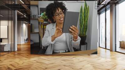 An african american woman with curly hair, sitting in an office, smiling and looking at a tablet, surrounded by indoor plants and shelves with books and files. Wall mural