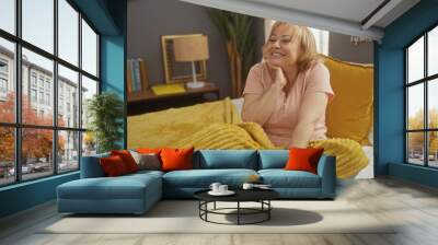 An elderly beautiful caucasian woman smiles joyfully while sitting on a cozy bed in a warmly lit bedroom at home. Wall mural