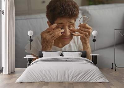 An elderly hispanic woman with short hair sits in a living room, rubbing her eyes while holding glasses in her hand. Wall mural