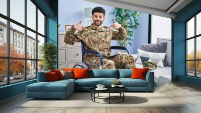 Arab man wearing camouflage army uniform sitting on wheelchair looking confident with smile on face, pointing oneself with fingers proud and happy. Wall mural