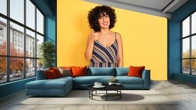 Arab woman with curly hair wearing striped colorful dress over isolated yellow background doing happy thumbs up gesture with hand. Approving expression looking at the camera showing success. Wall mural