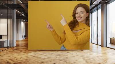 Attractive young caucasian woman with blonde hair smiling and pointing upward in a yellow sweater against a yellow background. Wall mural