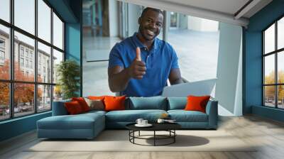 Attractive young man in a workplace giving a thumbs up while working on a laptop indoors. Wall mural